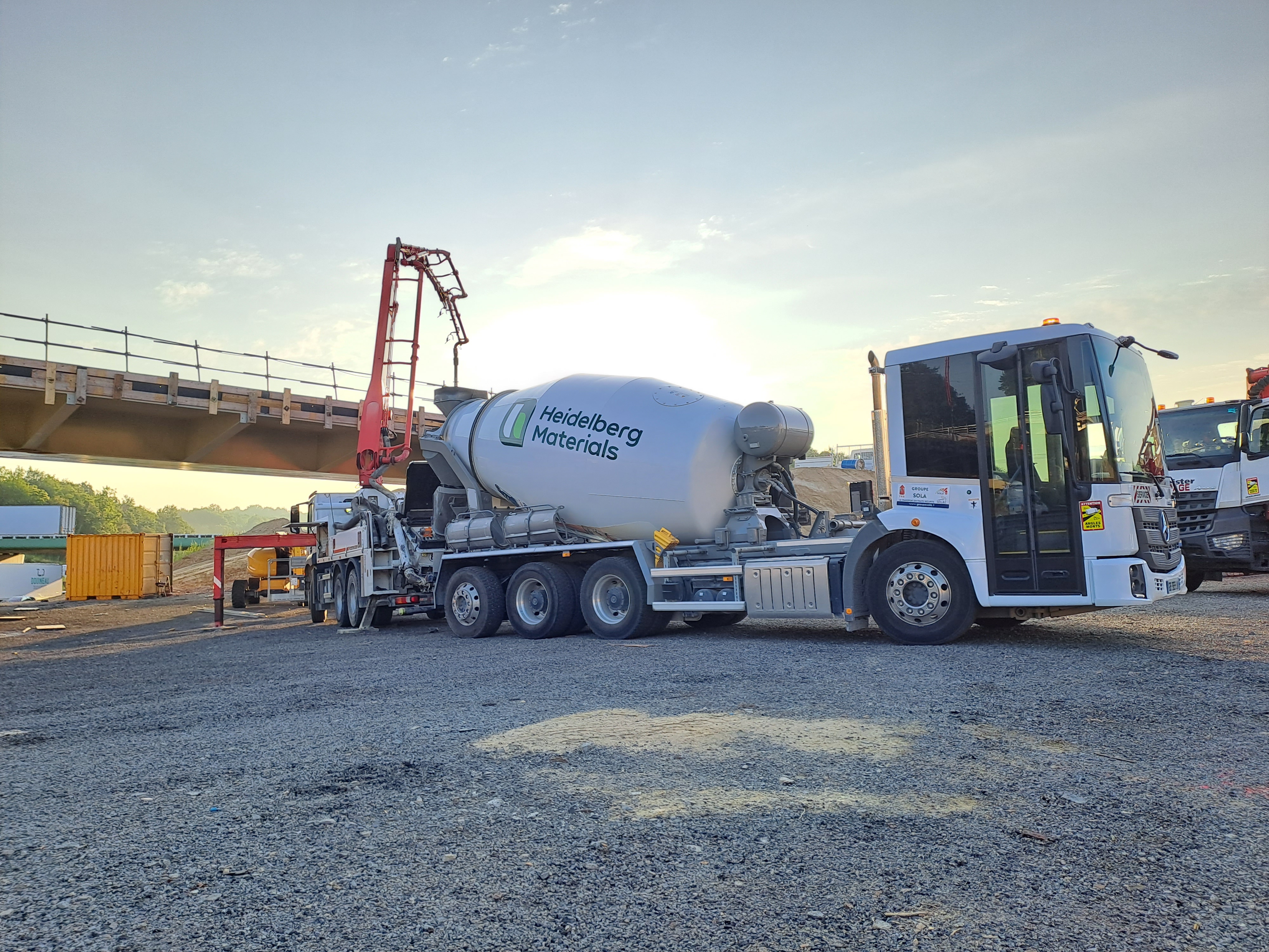 Camion-toupie blanc avec logo "Heidelberg Materials" stationné sur un chantier de construction. Une pompe à béton rouge est connectée au camion. En arrière-plan, un pont en construction et des conteneurs jaunes. Le ciel est dégagé avec une lumière matinale.