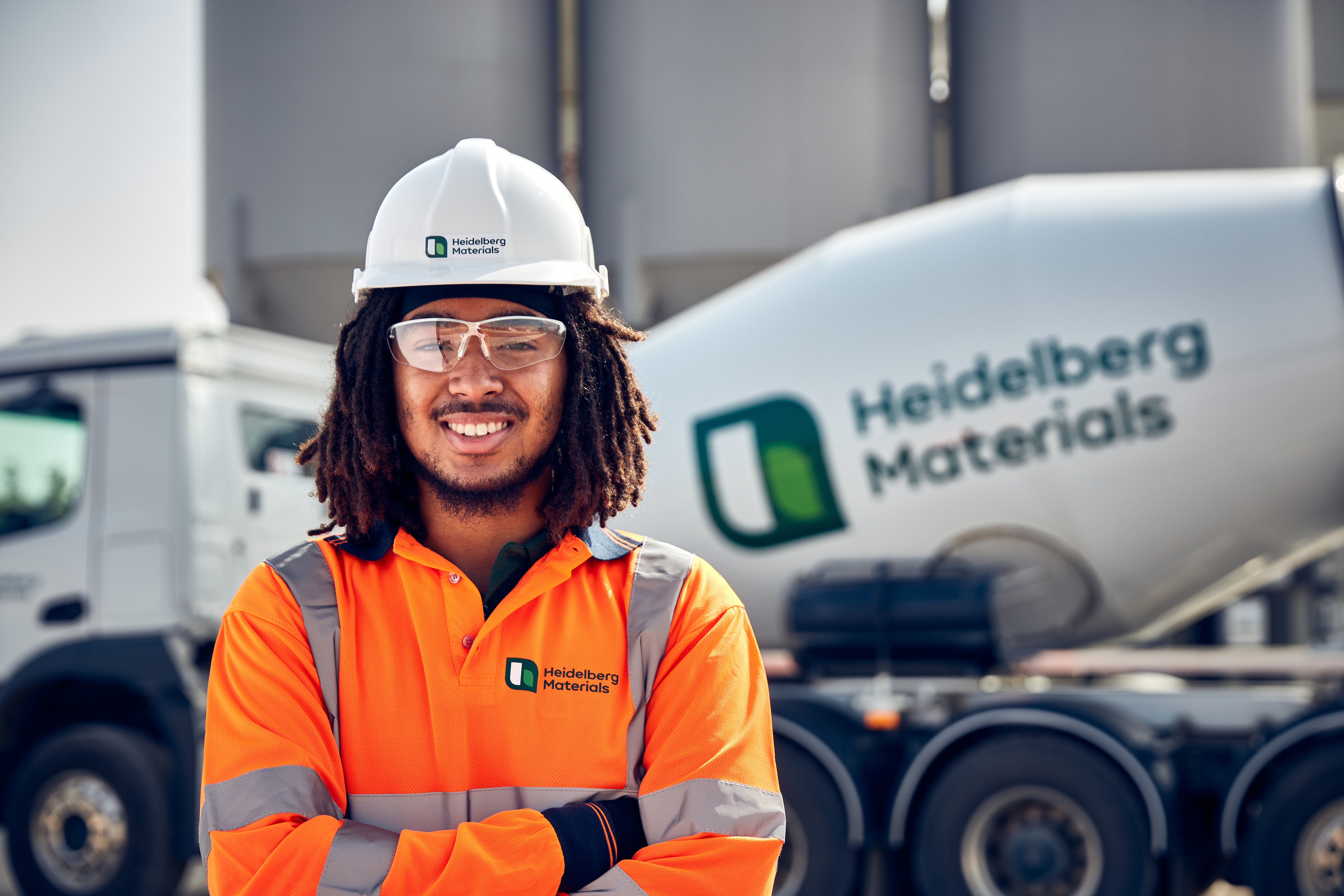 Employé de Heidelberg Materials en tenue de travail orange avec gilet réfléchissant et casque de sécurité blanc, debout devant une bétonnière arborant le logo de l’entreprise.