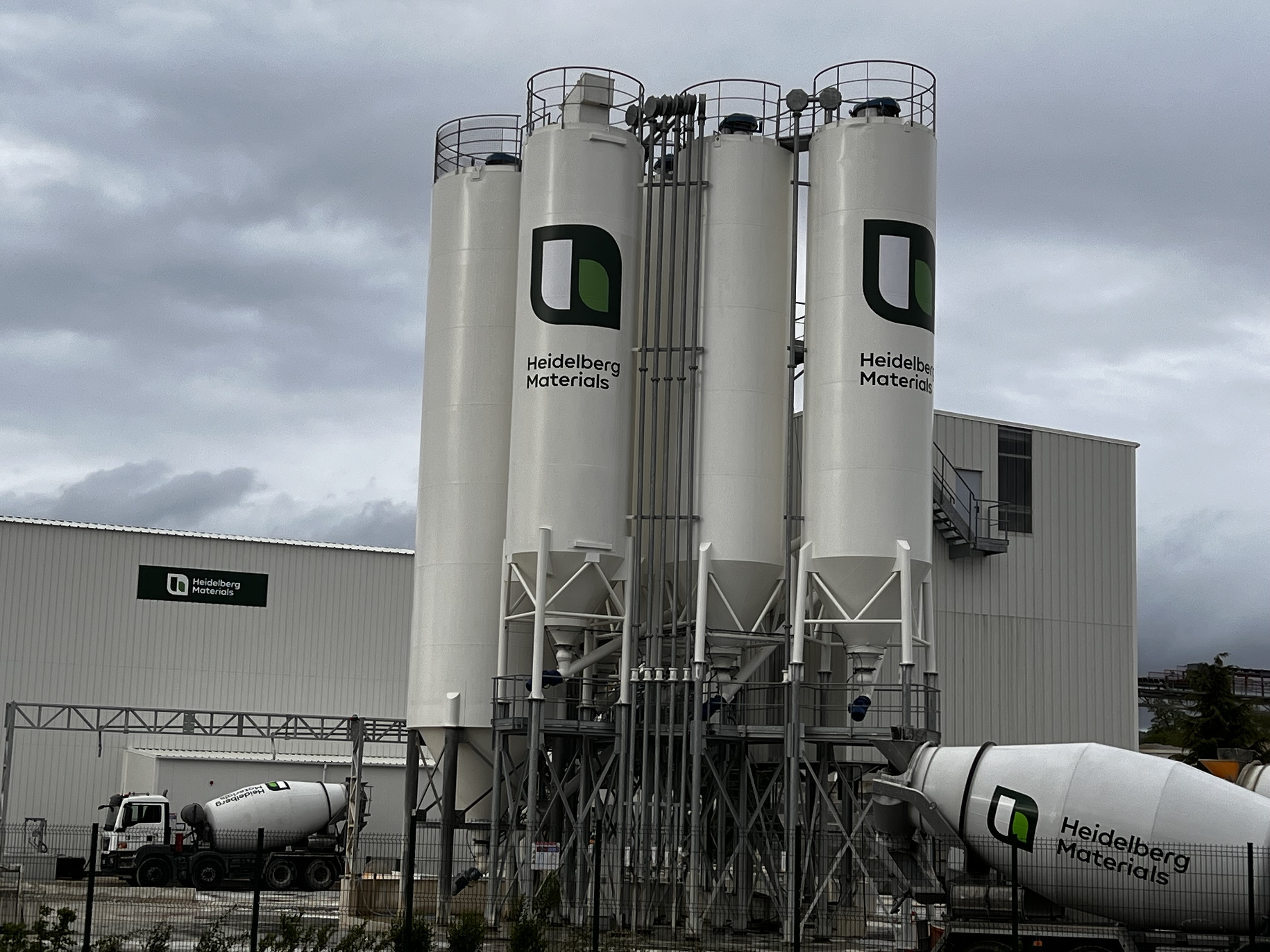 Usine de production de béton avec quatre grands silos blancs verticaux portant le logo « Lambe », un bâtiment industriel en arrière-plan, et deux camions-malaxeurs stationnés, l'un à gauche et l'autre à droite. Le ciel est nuageux.