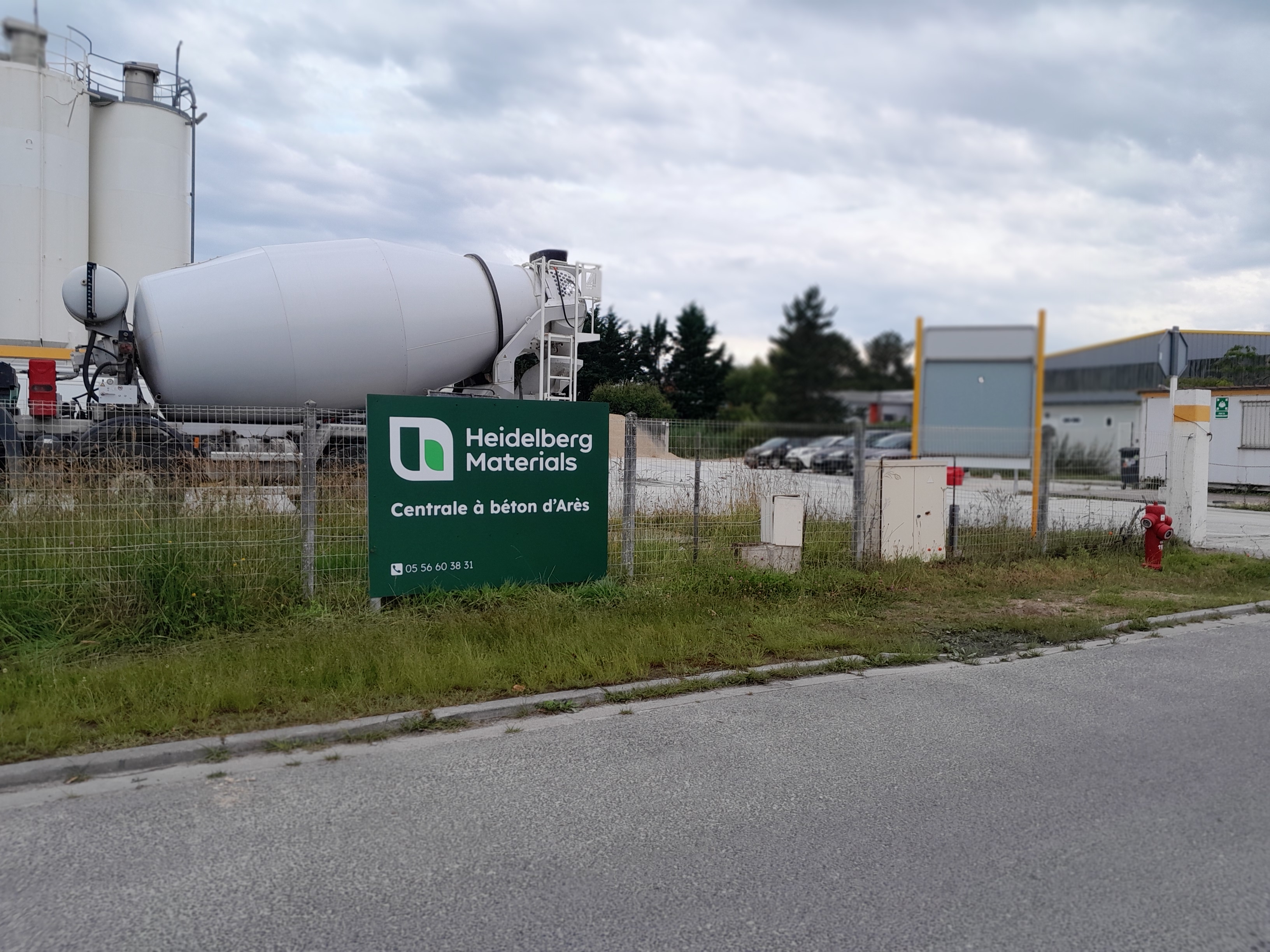 Usine Heidelberg Materials avec un silo blanc à béton à gauche, panneau vert au premier plan indiquant "Construire le futur d'ici", clôture métallique entourant le site, et route asphaltée au bas de l'image.