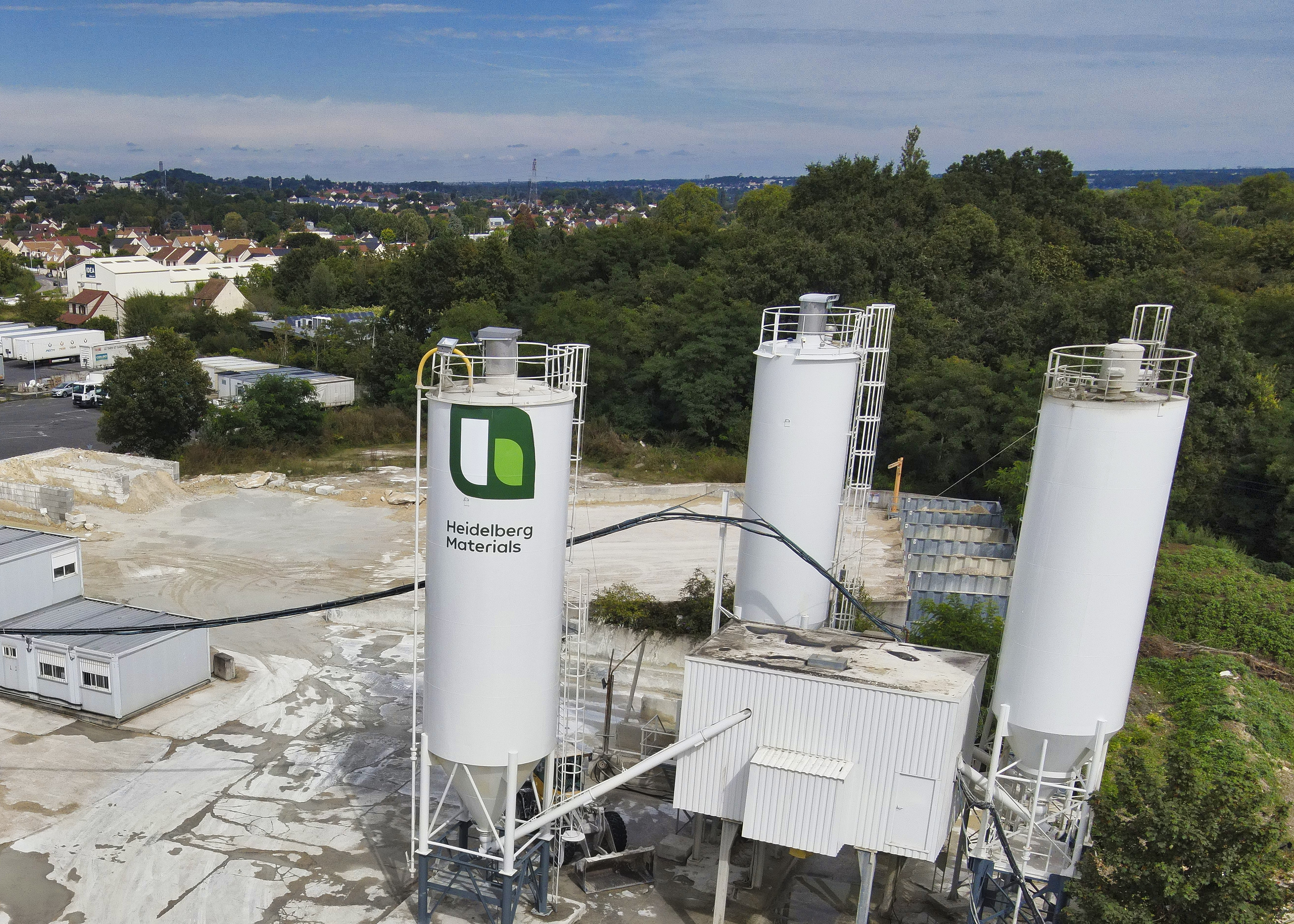 Vue aérienne d'une station de béton avec trois silos blancs, dont l'un porte le logo vert et noir d'une entreprise. Le site est entouré de végétation dense à droite et d'habitations en arrière-plan à gauche. Structures métalliques reliant les silos visibles au centre.