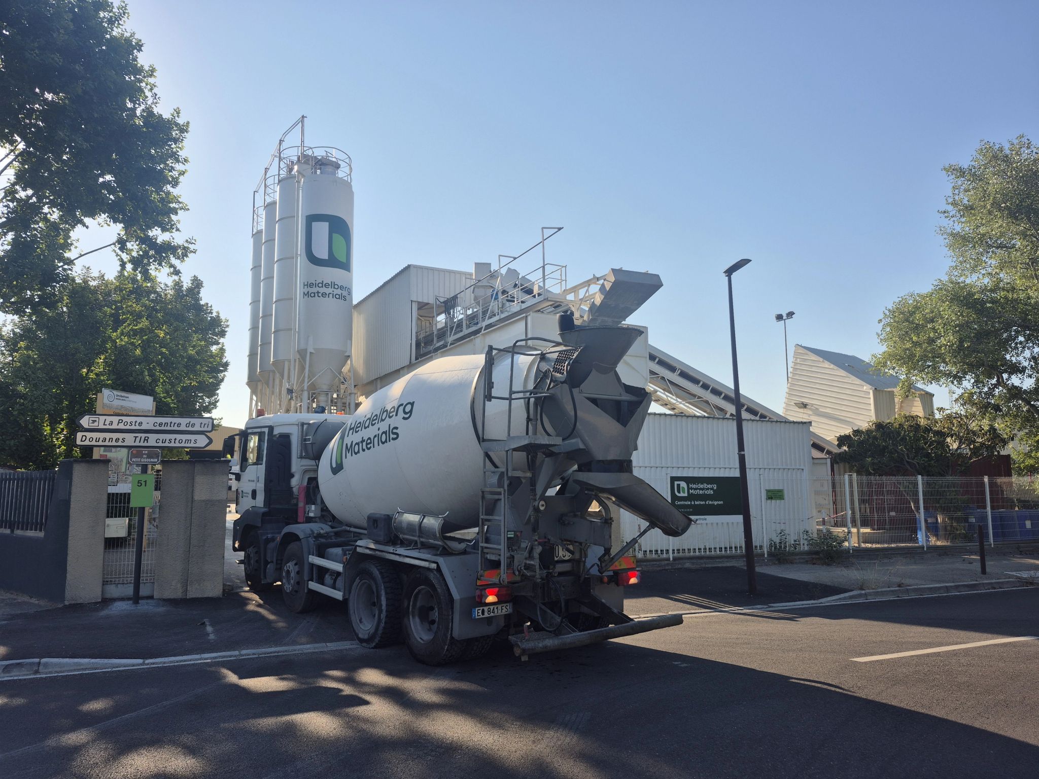 Camion-toupie de béton garé devant une centrale à béton, avec un silo blanc marqué d’un logo vert et entouré de structures industrielles. La scène est éclairée par le soleil, avec des arbres en arrière-plan et une clôture métallique délimitant le site.