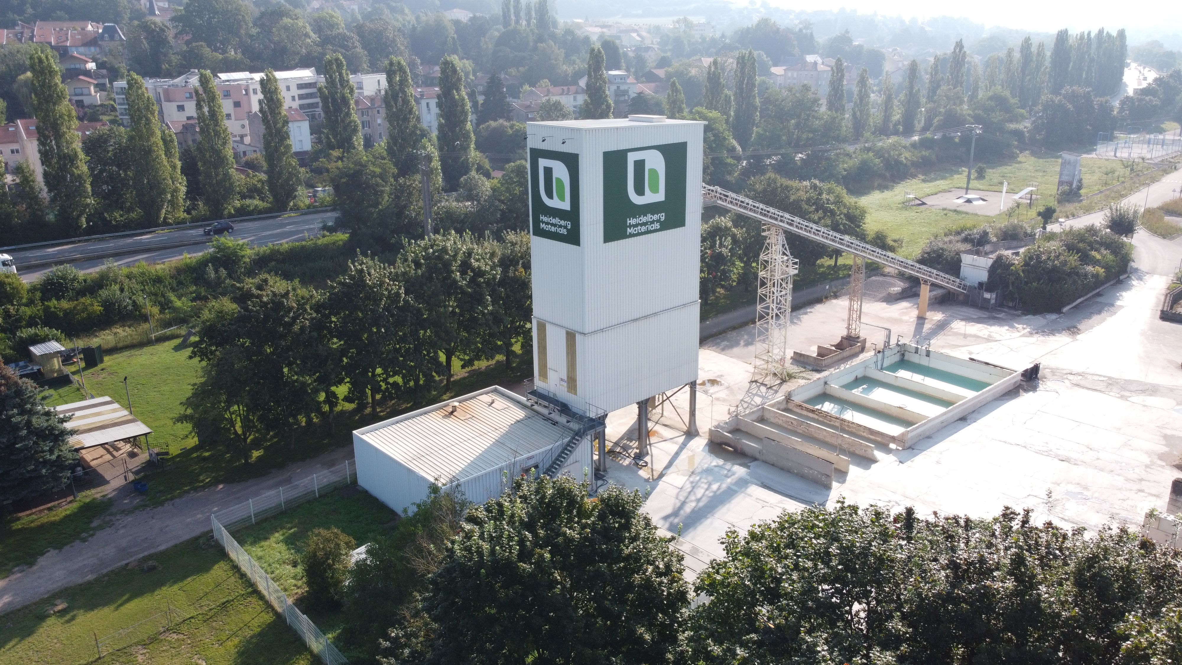 Vue aérienne d’une centrale à béton moderne avec une grande tour blanche au centre, ornée du logo de l’entreprise. Des espaces de stockage et des installations métalliques sont situés à droite. En arrière-plan, une route bordée d'arbres mène à une zone résidentielle.