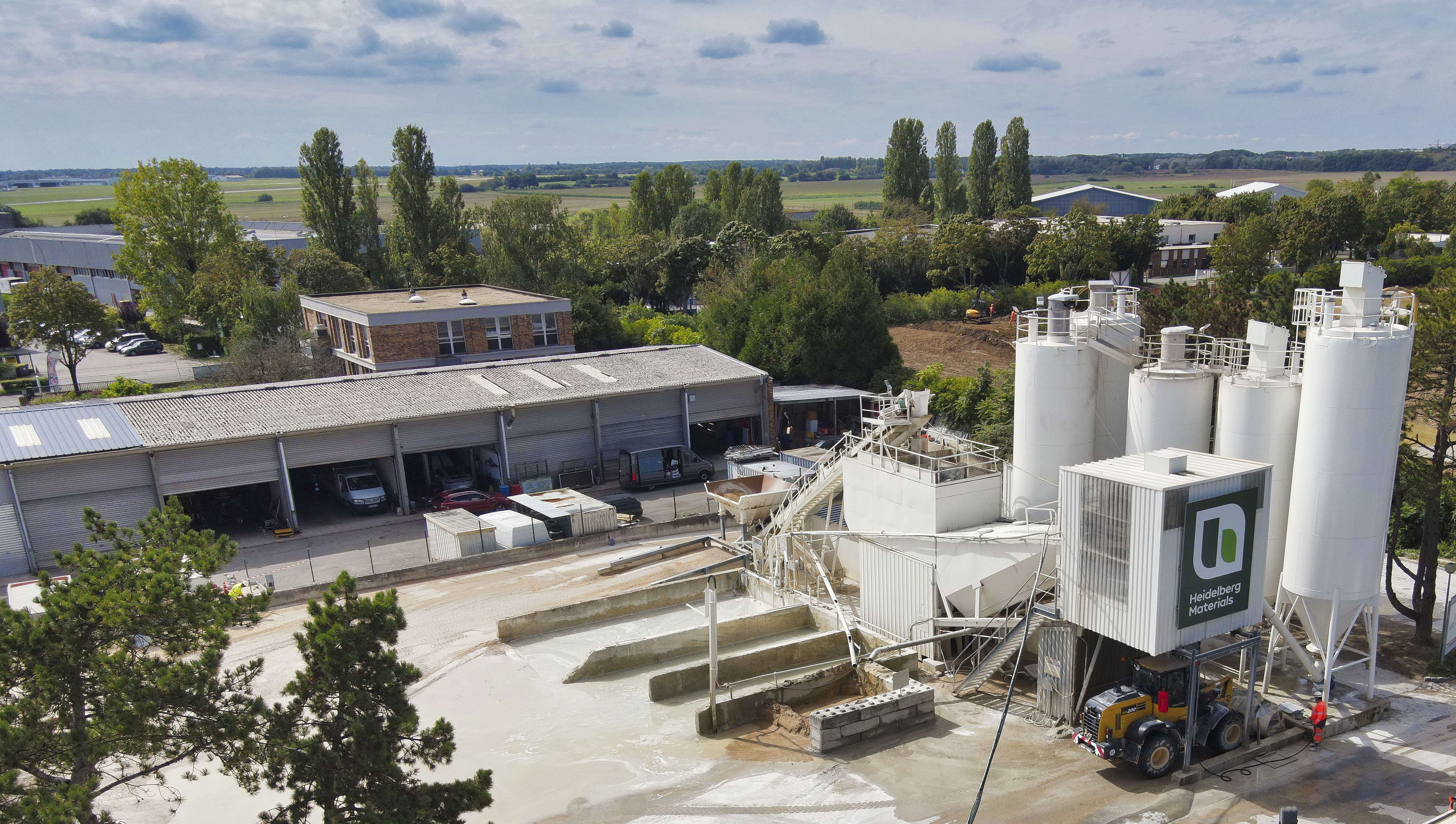 Vue aérienne d'une usine de production de béton, comprenant des silos blancs verticaux à droite avec le logo visible sur l'un d'eux. À gauche, des bâtiments industriels en enfilade et des zones de stockage. En arrière-plan, paysage rural avec arbres et champs sous un ciel partiellement nuageux.