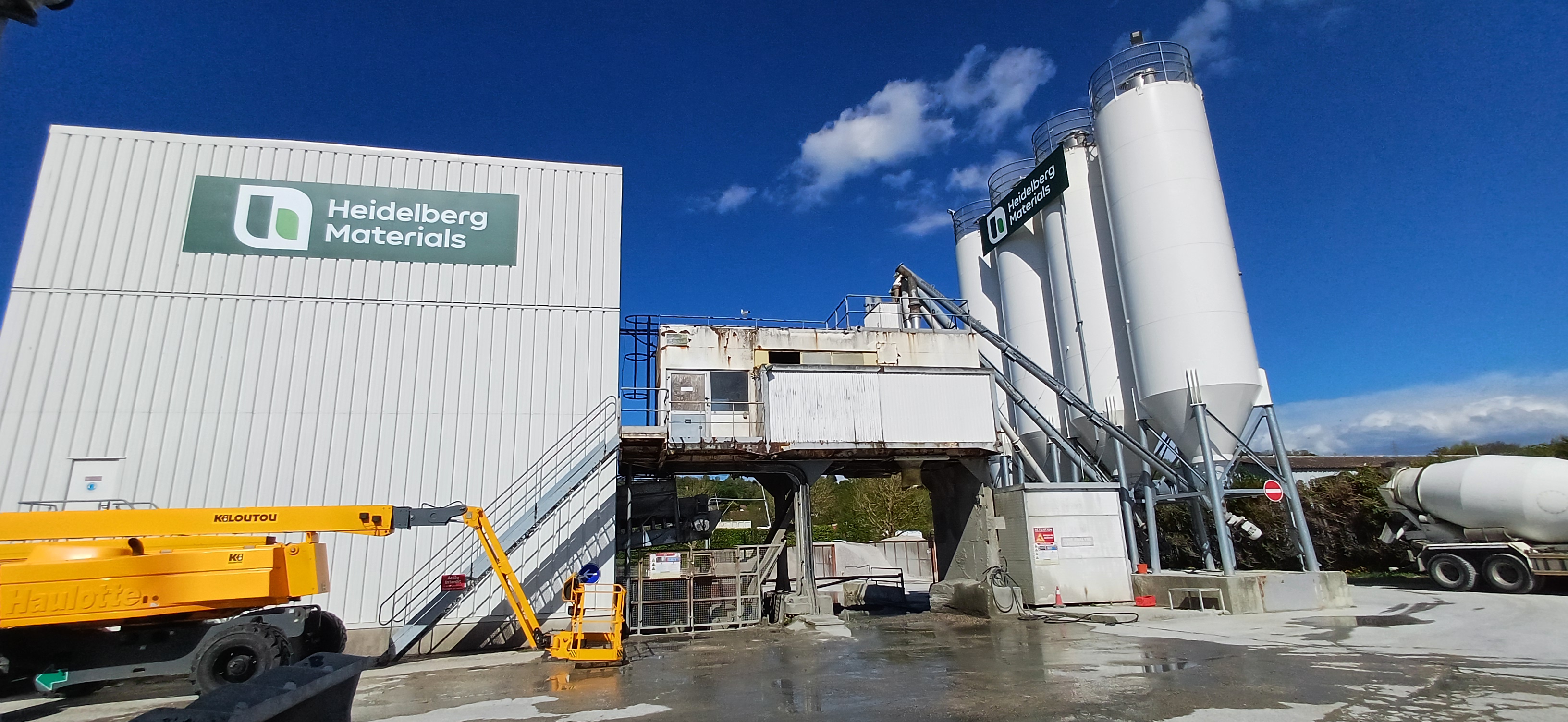 Usine de production de béton Heidelberg Materials en extérieur avec un bâtiment blanc sur la gauche, plusieurs silos verticaux blancs sur la droite, et un camion malaxeur garé près des silos. Une grue jaune est visible au premier plan gauche, ciel bleu en arrière-plan.