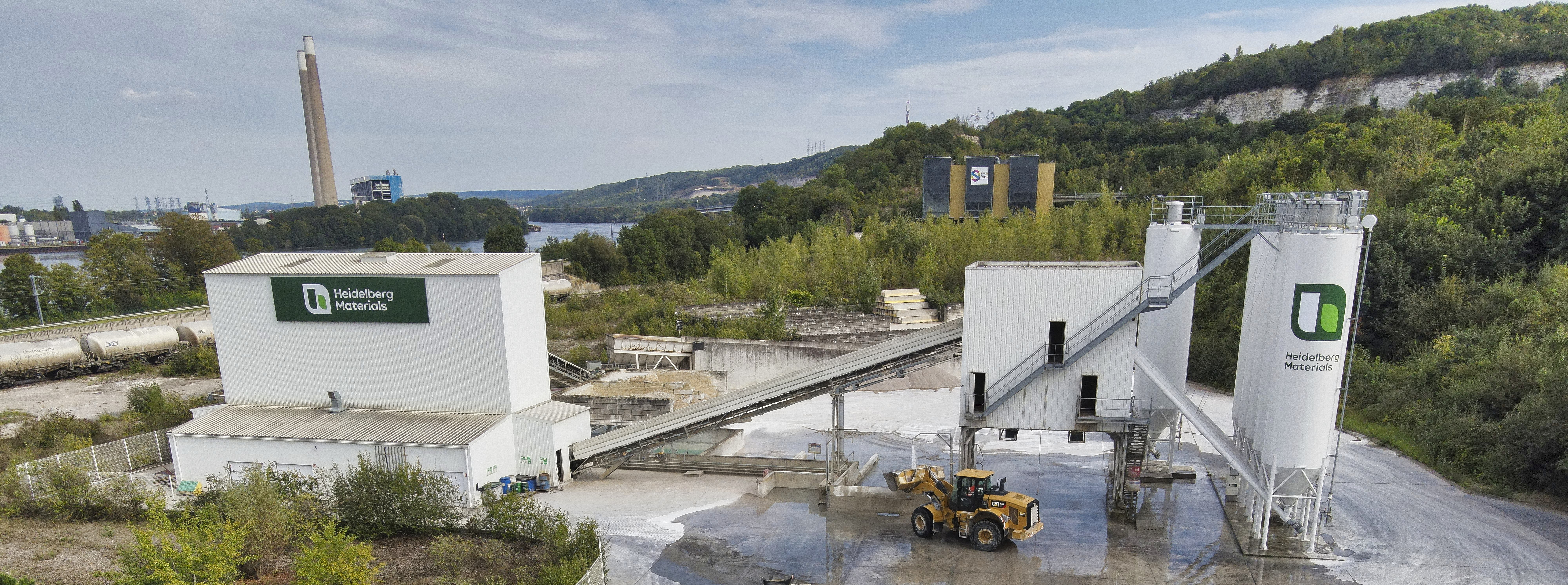 Usine de production de béton située en extérieur, avec deux grands silos blancs à droite, un bâtiment principal blanc à gauche portant le logo de l’entreprise, et une cour centrale équipée de convoyeurs et d’un chargeur sur pneus jaune. En arrière-plan, des collines verdoyantes et des bâtiments industriels.