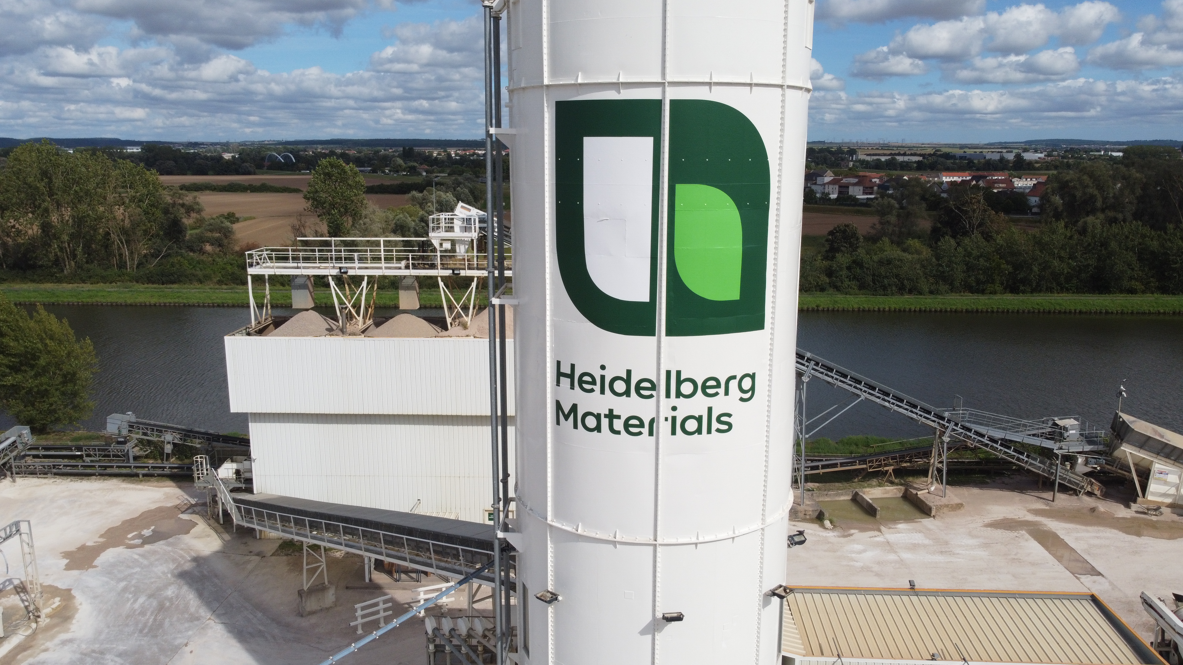 Tour de stockage blanche avec le logo de Heidelberg Materials, affichant des formes vertes et blanches abstraites. À l’arrière-plan, un cours d’eau entouré de champs et de végétation, ainsi que des équipements industriels tels que des convoyeurs visibles en bas à gauche. Ciel partiellement nuageux.