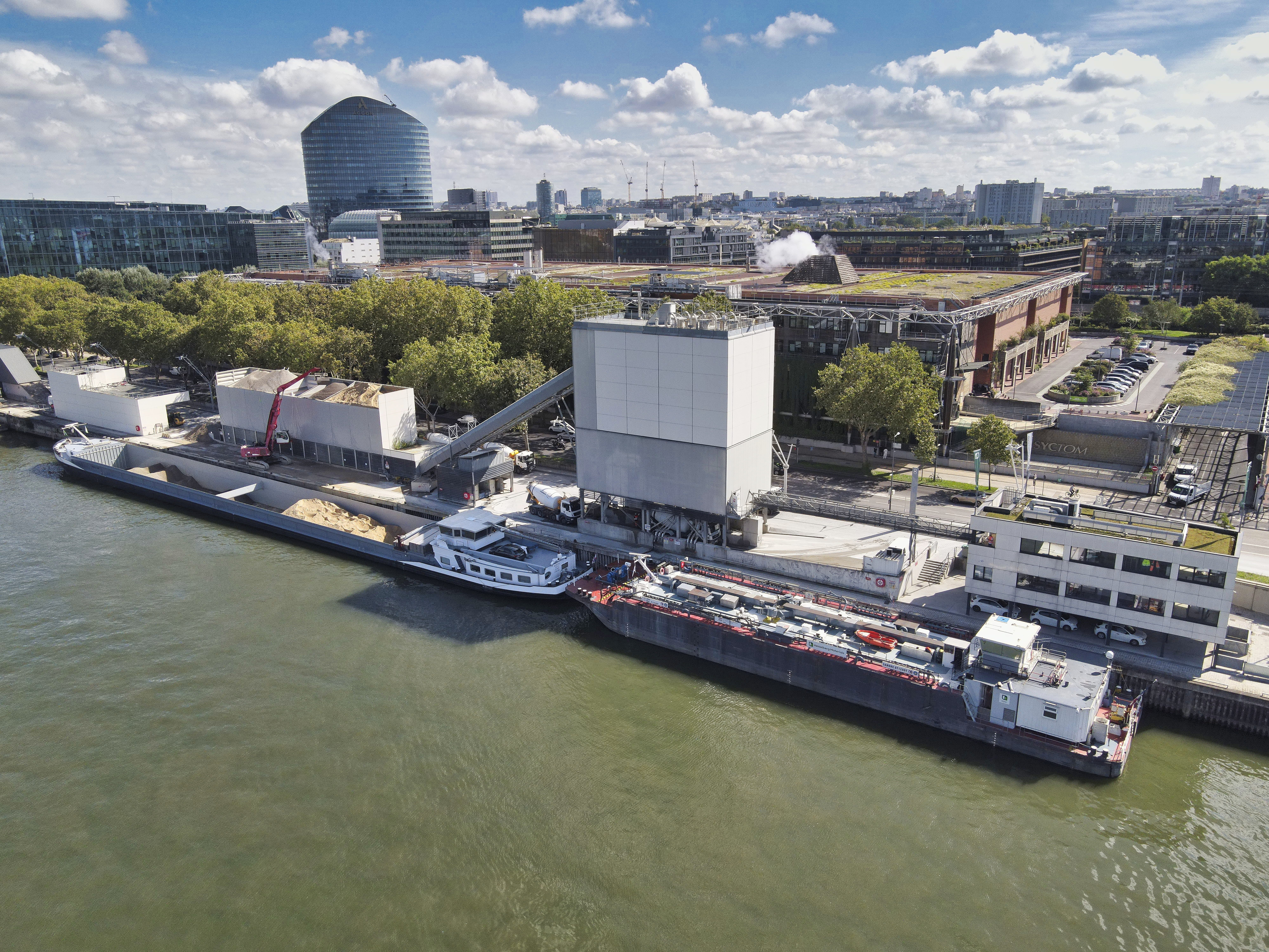 Vue aérienne d’un site portuaire au bord d’un fleuve, avec des bâtiments industriels modernes, des péniches amarrées le long du quai et des espaces verts en arrière-plan. Le ciel est dégagé, et des bâtiments urbains et une grande structure arrondie apparaissent à l’horizon.