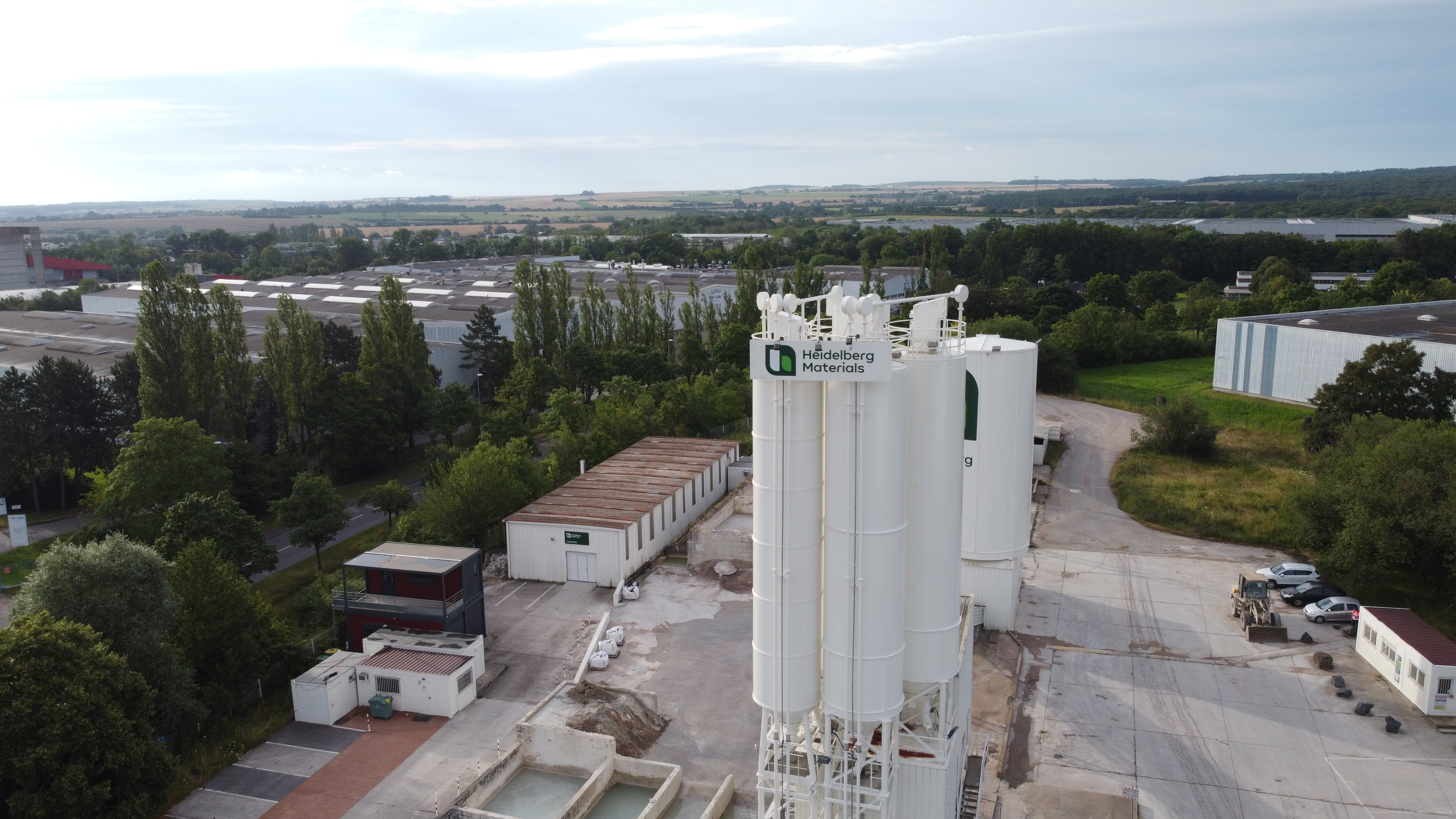 Vue aérienne d'un site industriel avec plusieurs silos blancs centraux portant un logo vert, entourés de bâtiments et d'arbres. À l'arrière-plan, un paysage semi-urbain avec des champs et des infrastructures.