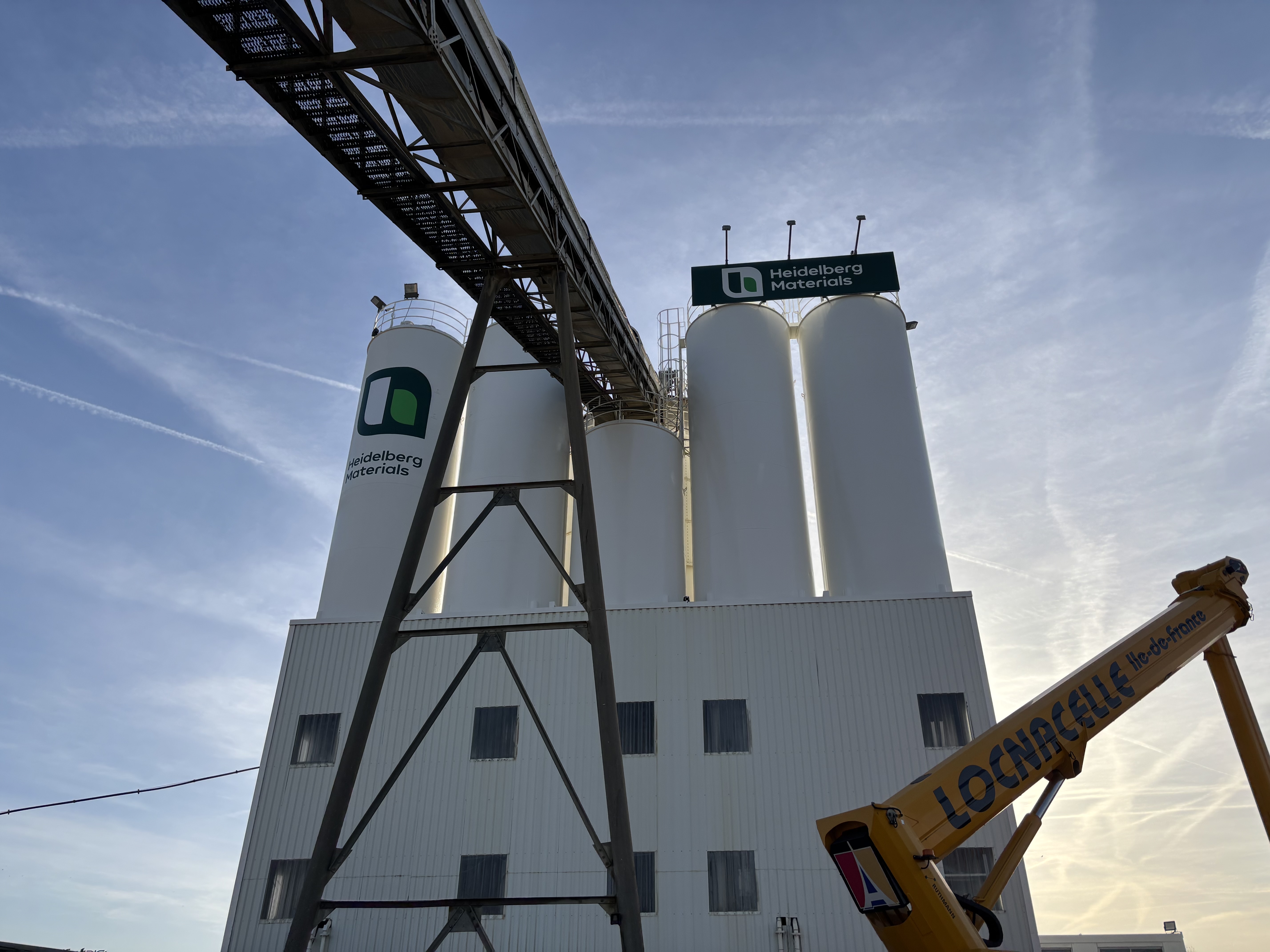 Vue en contre-plongée d'une usine composée de quatre grands silos blancs avec le logo de la société Unibéton visible sur deux d'entre eux. Une structure métallique pour un convoyeur traverse en diagonale le haut de l'image. En bas à droite, le bras d'une grue jaune marquée "LOCHBIELI". Ciel clair avec des traînées de condensation en arrière-plan.