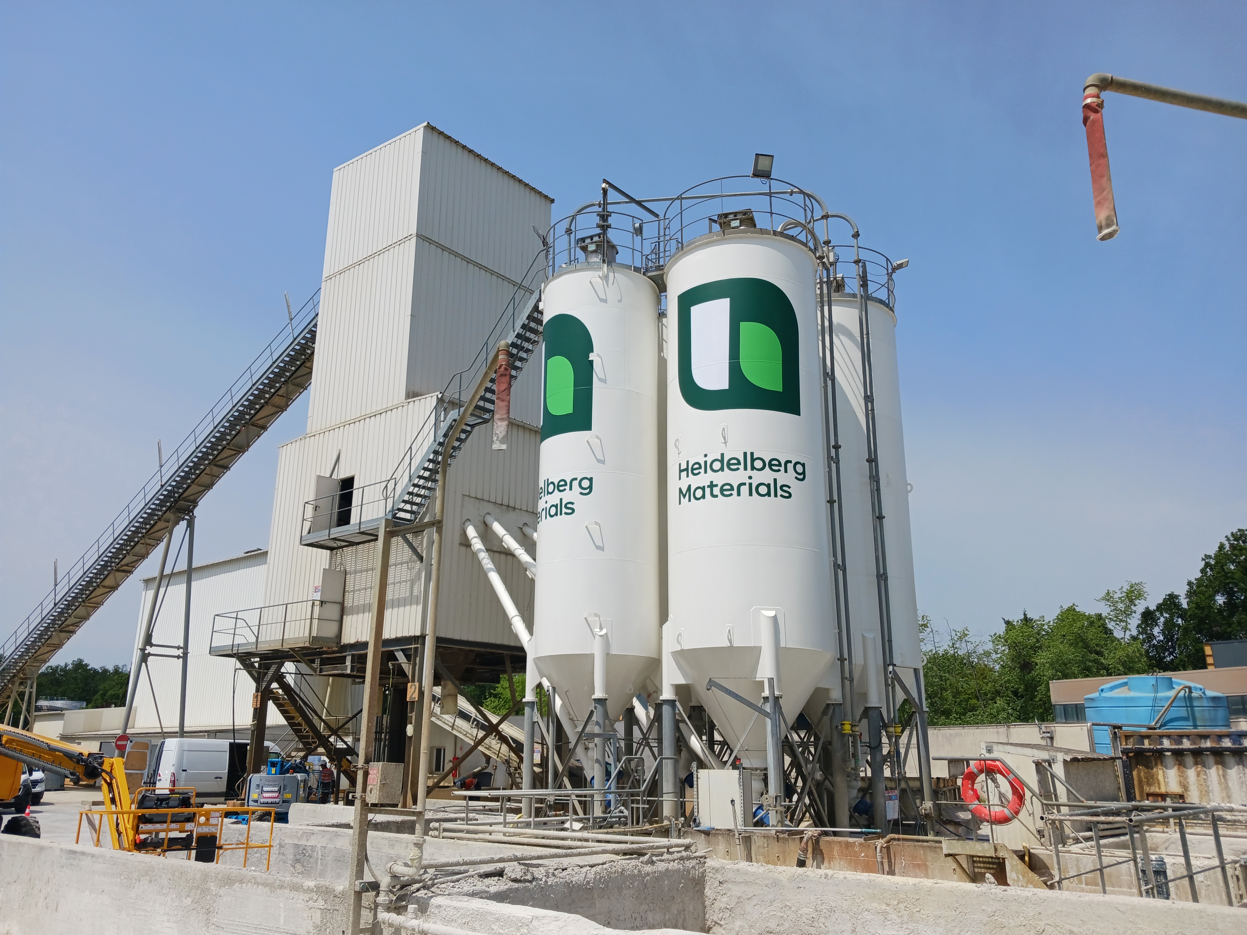 Deux silos blancs avec le logo « Heidelberg Materials » au centre d'une centrale à béton, entourés de structures métalliques et d’équipements industriels. Une grue et des arbres sont visibles à droite en arrière-plan, sous un ciel bleu.