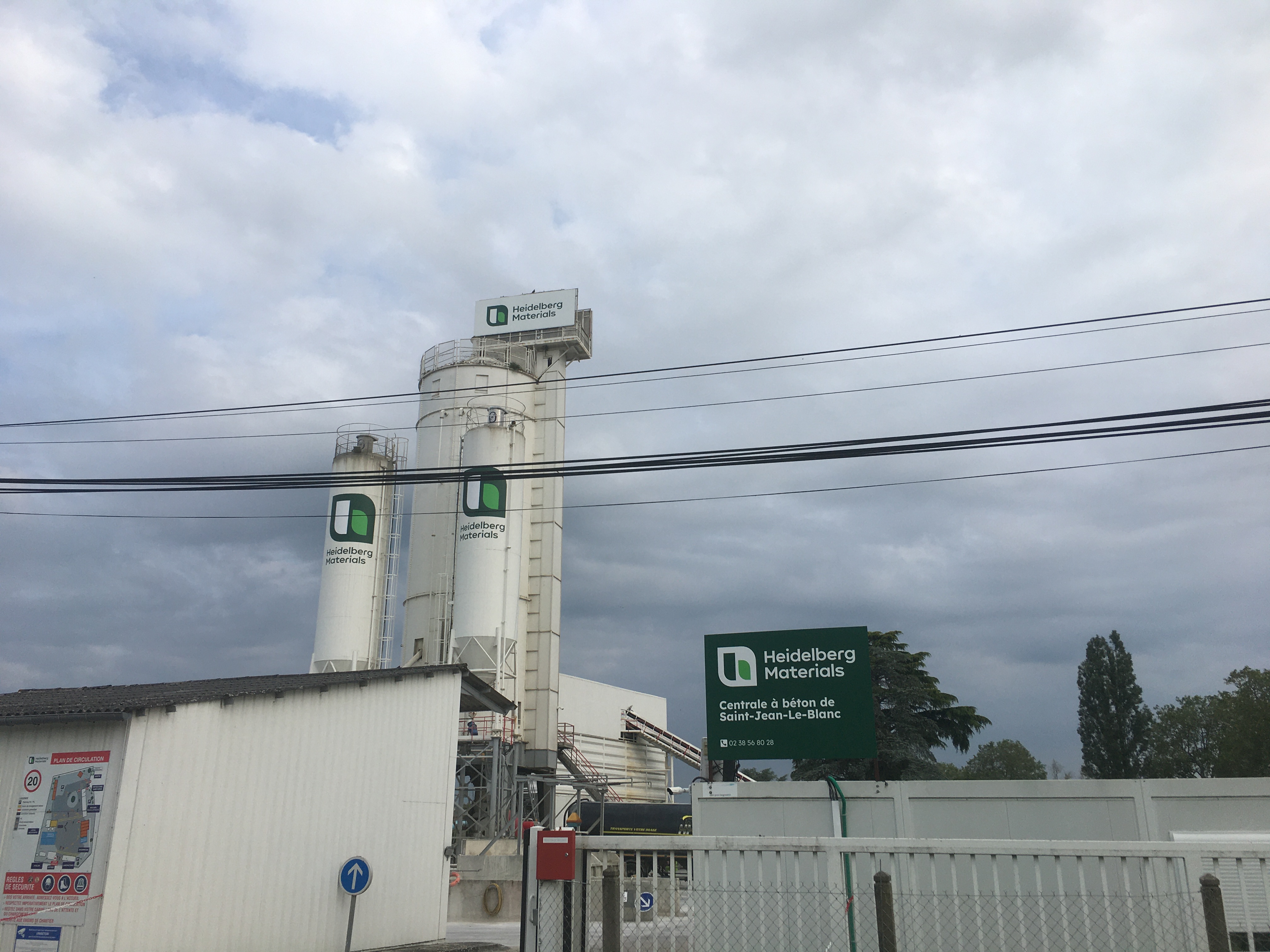 Deux silos blancs de Heidelberg Materials avec logo, entourés de clôtures et structures industrielles, sous un ciel nuageux. Un panneau vert à droite indique le nom de l’entreprise.