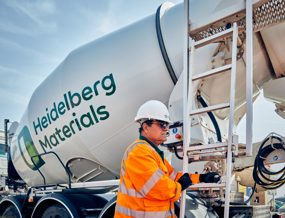 Employé en gilet orange et casque blanc debout près d'un camion-toupie marqué "Heidelberg Materials", tenant une échelle métallique, avec ciel dégagé en arrière-plan.