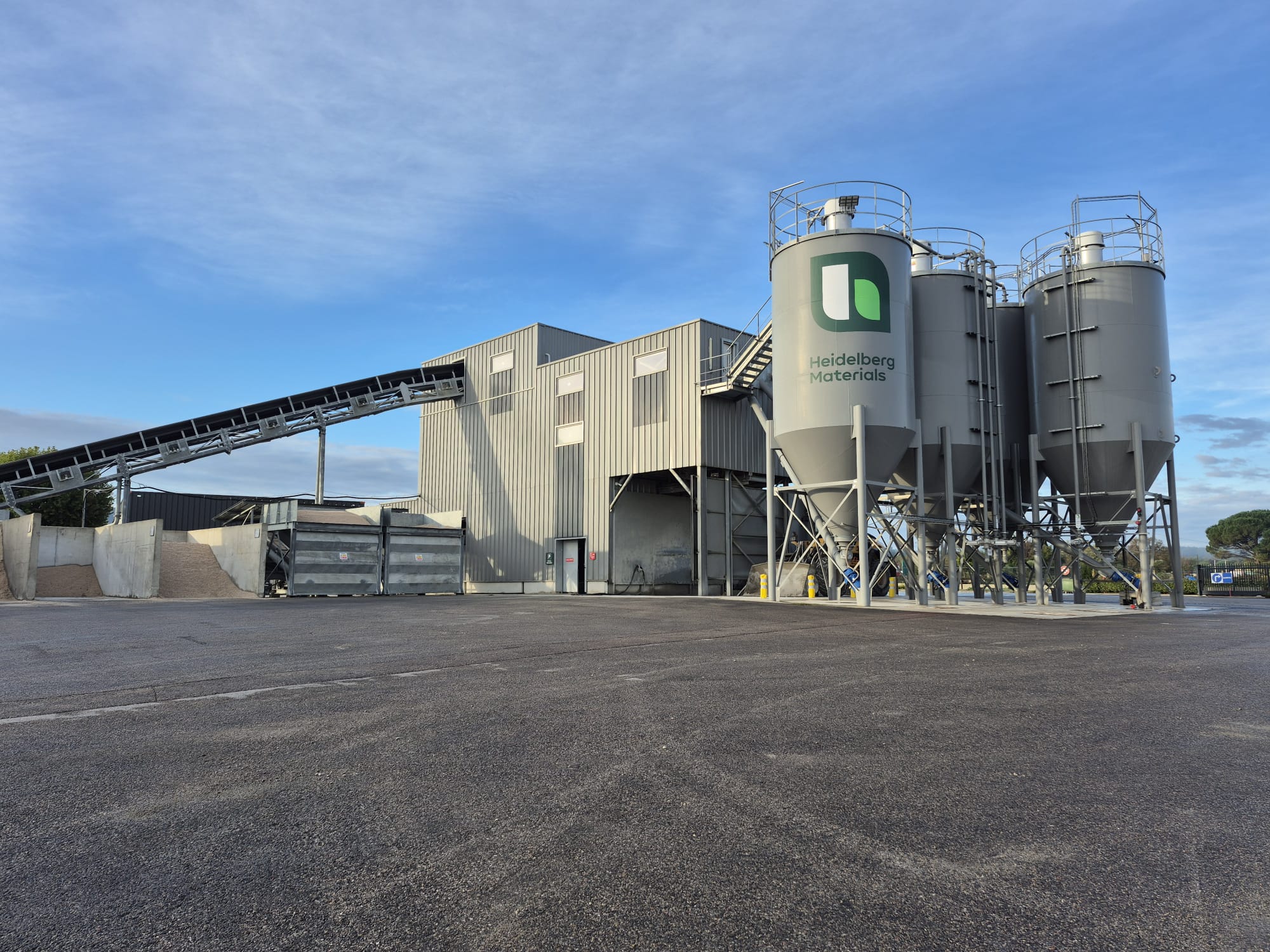 Installation industrielle avec plusieurs silos métalliques à droite, un convoyeur incliné à gauche, et un bâtiment central gris. Le sol est asphalté et le ciel est dégagé avec quelques nuages. Logo vert sur les silos.