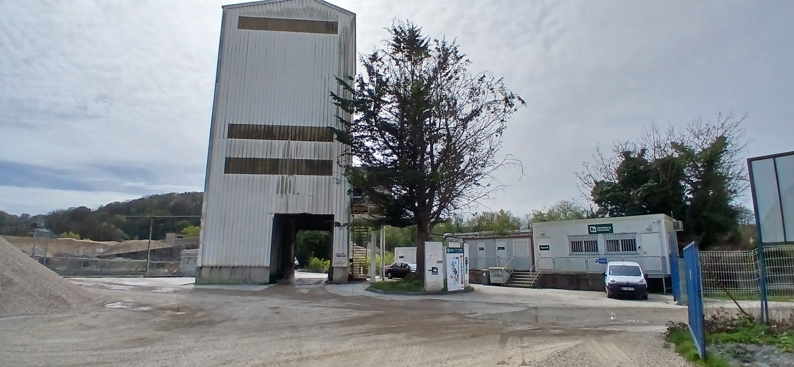 Site industriel avec un bâtiment principal en béton, entouré d'arbres et de petites structures annexes.
