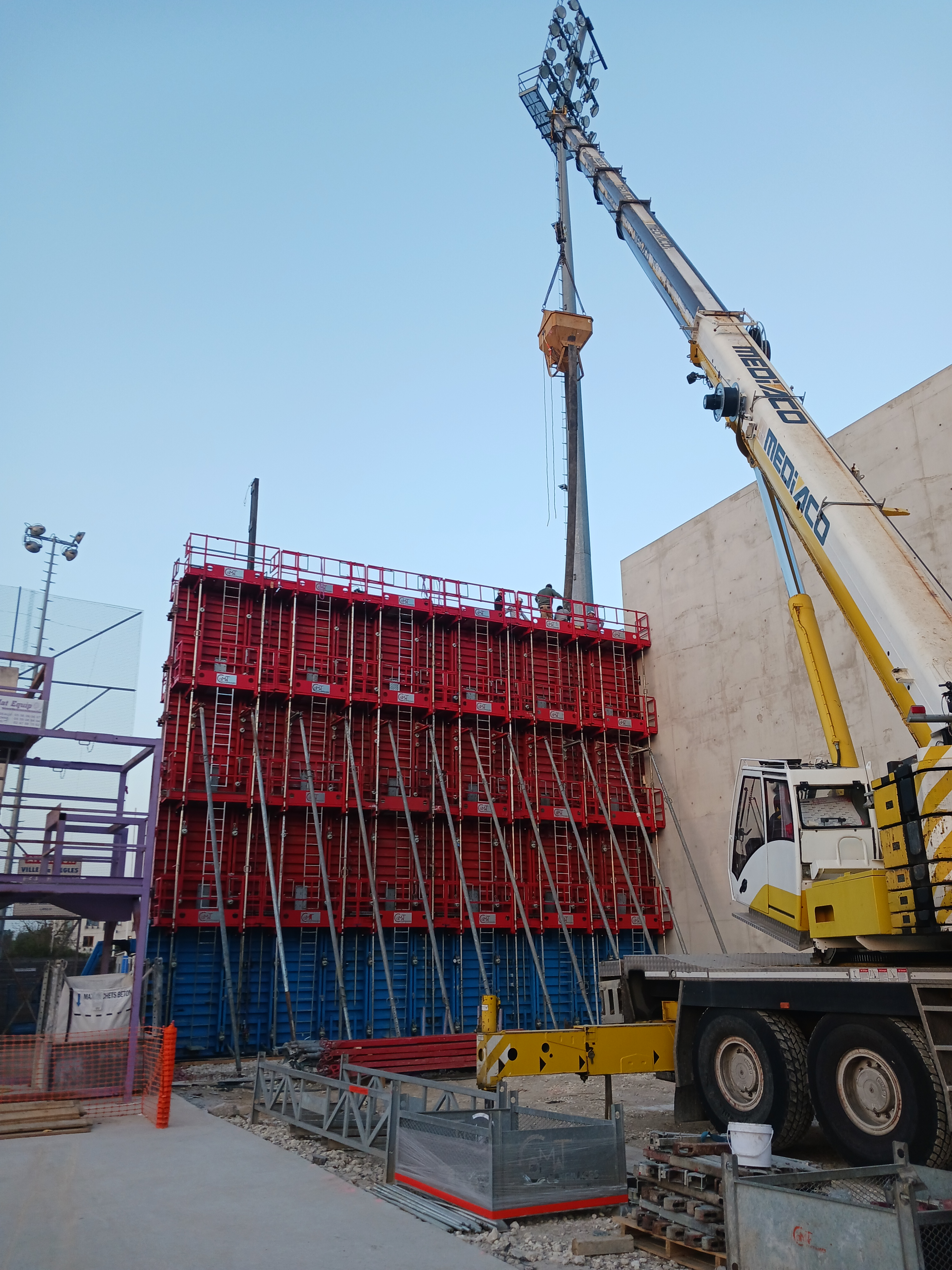 Grue soulevant du matériel de construction sur un chantier avec des coffrages en béton rouge et bleu.