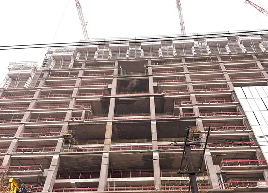 Immeuble en construction vu en contre-plongée, avec une structure en béton et plusieurs étages en cours de finition. Des grues sont visibles en haut, et un feuillage d'arbre se trouve en haut à gauche.