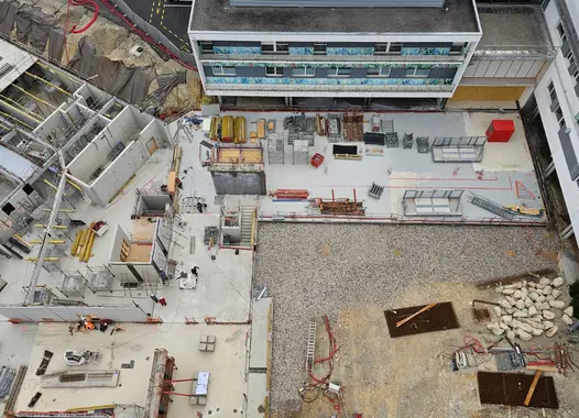 Vue aérienne d'un chantier de construction, montrant des structures bétonnées incomplètes à gauche, une zone centrale dégagée avec du gravier et des matériaux de construction, une excavation au bas à droite, et un bâtiment moderne achevé en haut. Des équipements de chantier et des zones de travail balisées sont visibles.